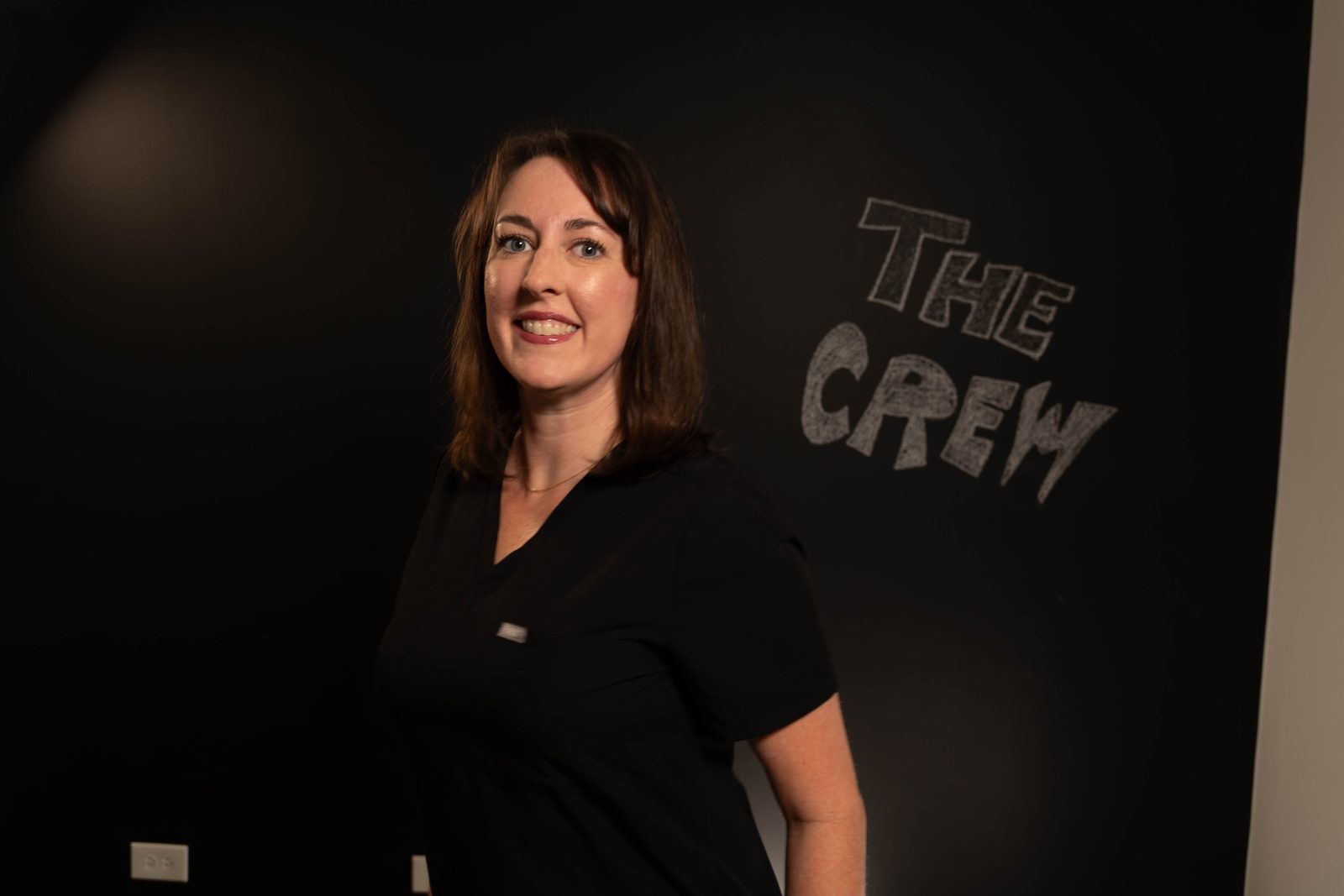 Smiling person in black scrubs standing in front of a chalkboard wall