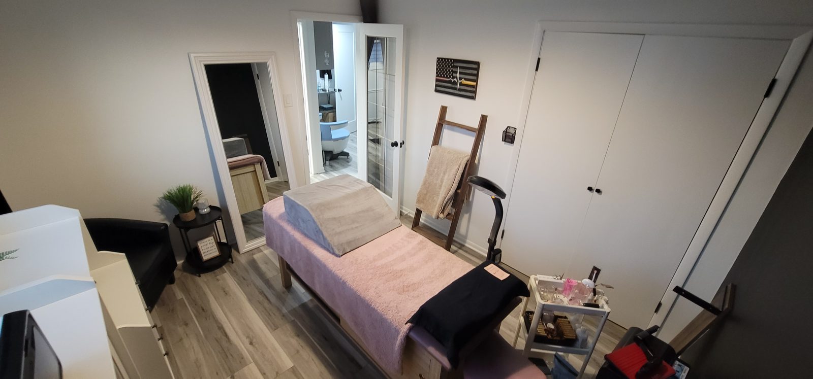 Wide view of a treatment room with a pink-blanketed treatment bed, a mirror and a ladder with a towel
