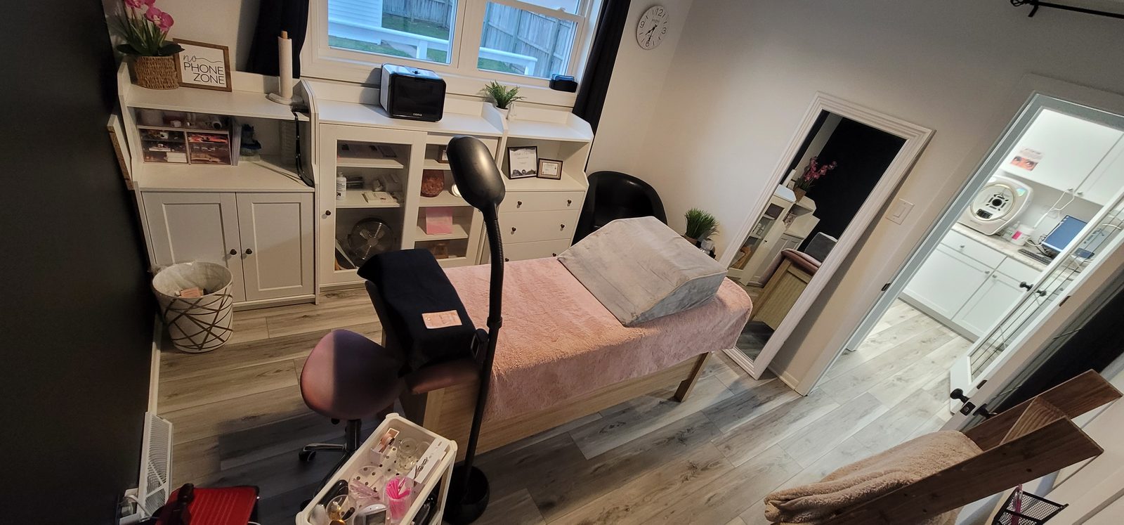 Treatment room with a pink-covered treatment bed, white cabinets, a magnifier lamp and natural light